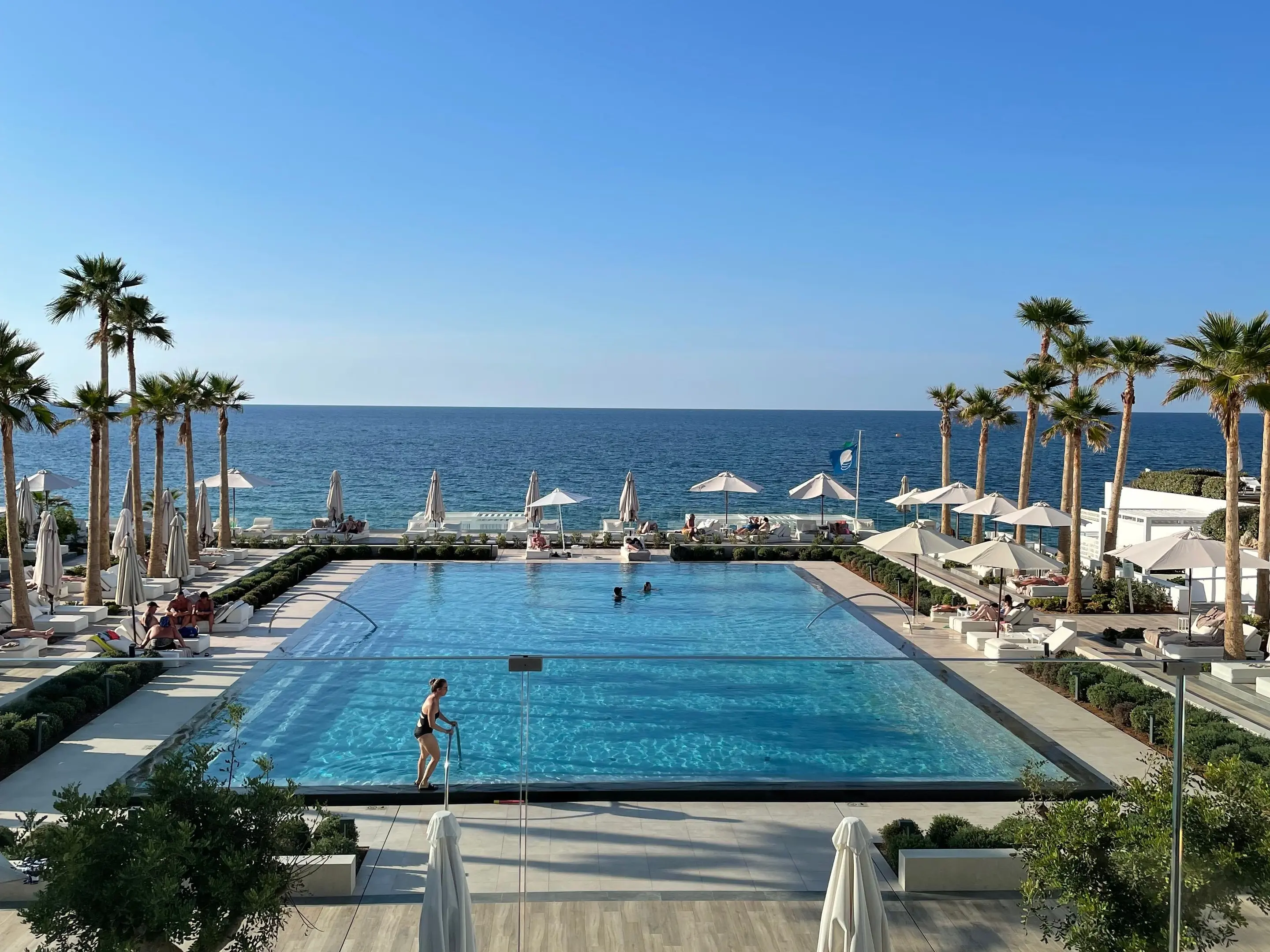 Piscina unui hotel de pe malul mării în Creta, cu șezlonguri albe și umbrele, într-o zi senină.