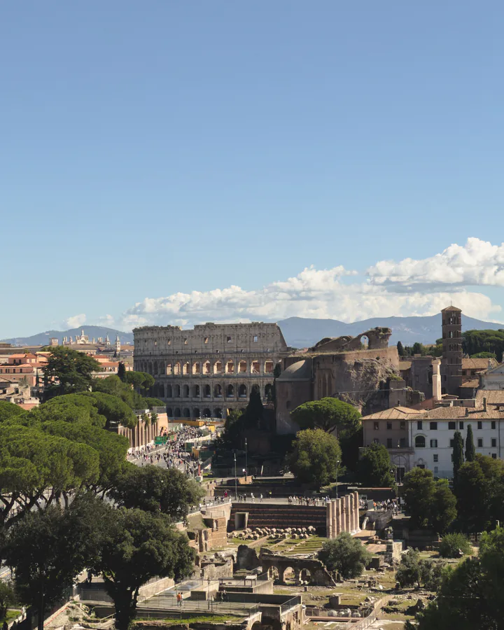 Roma văzută de sus: Colosseumul și Forul Roman între pini, cu munții în depărtare, sub cer albastru.