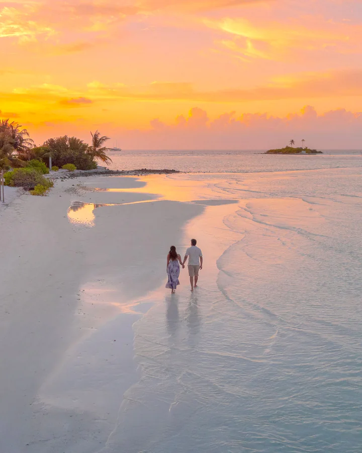 Un cuplu se plimbă pe o plajă albă din Maldive, la apus, cu vile pe apă în depărtare.