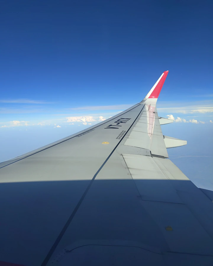 Aripa unui avion deasupra norilor, pe cer albastru, văzută de la geam.